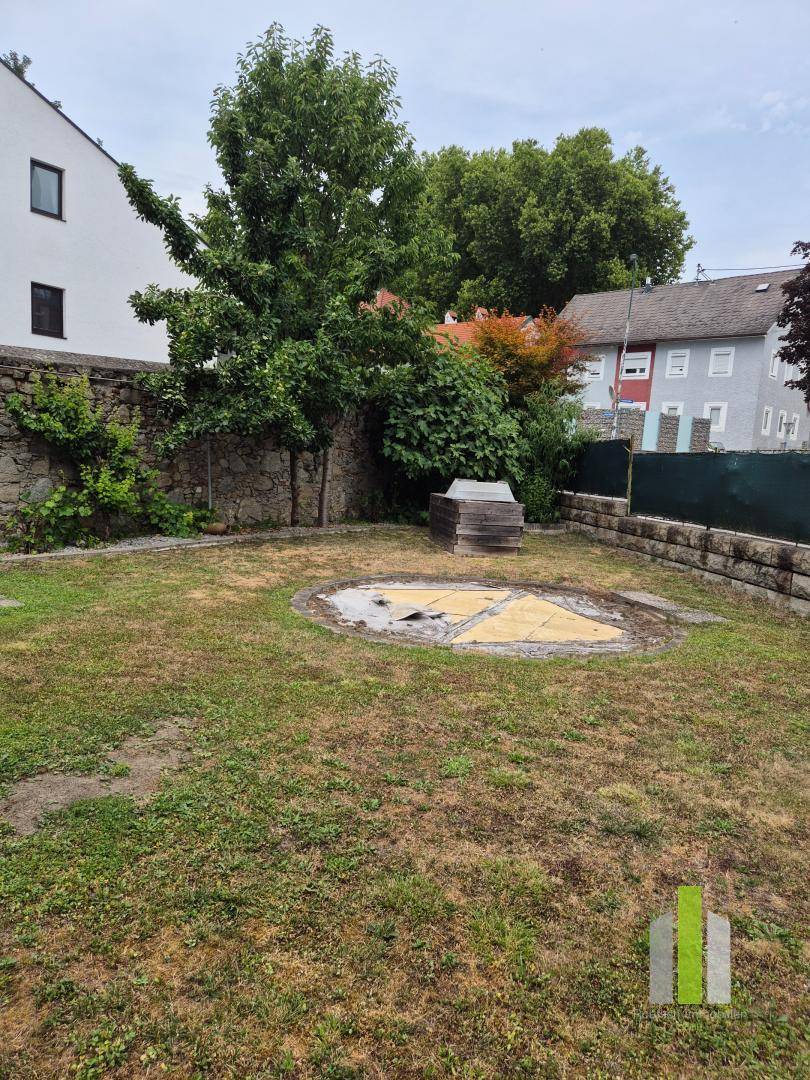 Gartenfläche mit Rasen, alten Bäumen und einer runden gepflasterten Fläche in der Mitte.