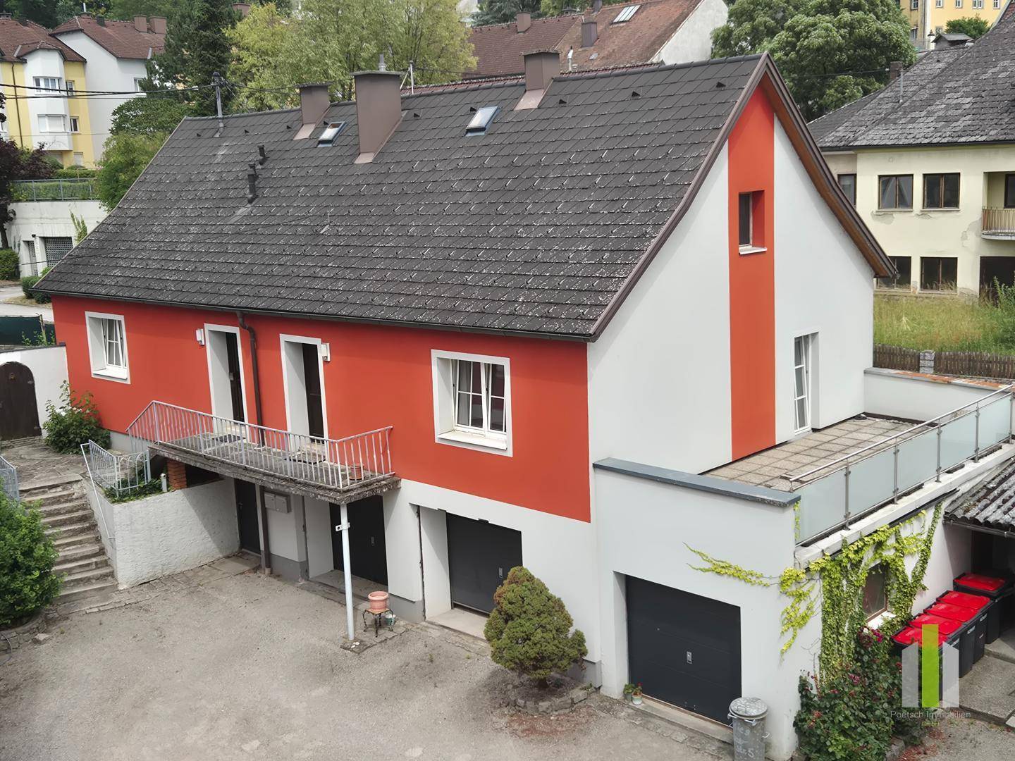 Rotes und weißes Haus mit Satteldach, Garagen und einem Balkon in ländlicher Umgebung.