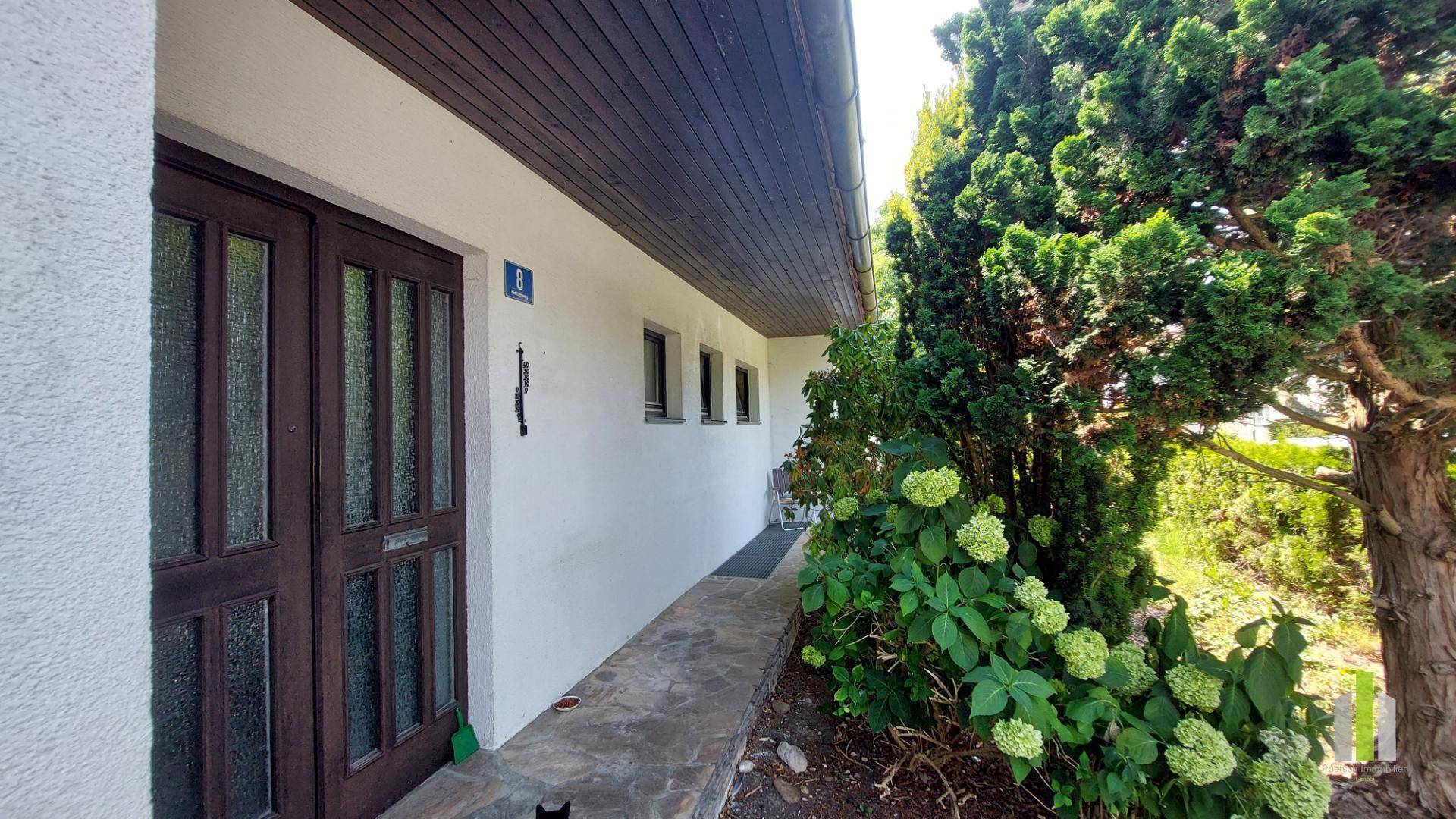 Eingangsbereich des Hauses mit Holztür und seitlichen Fenstern, umgeben von üppigem Grün.