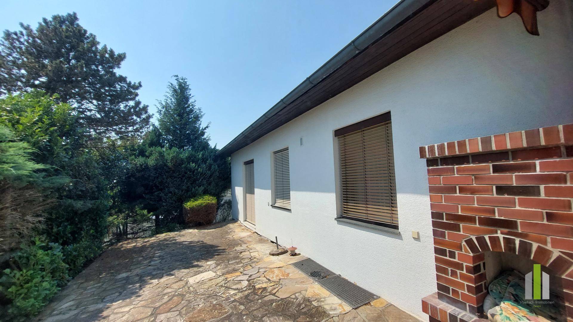 Seitliche Terrasse mit Steinplatten und gemauertem Grill, umgeben von grünen Büschen.