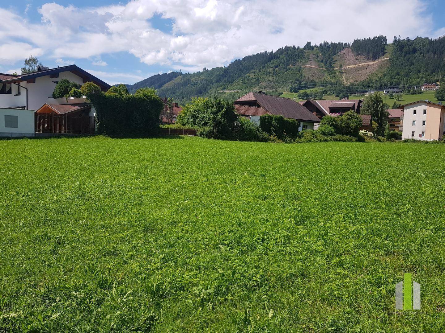 Weitläufiges grünes Feld mit Blick auf bewaldete Hügel und umliegende Wohnhäuser unter blauem Himmel.