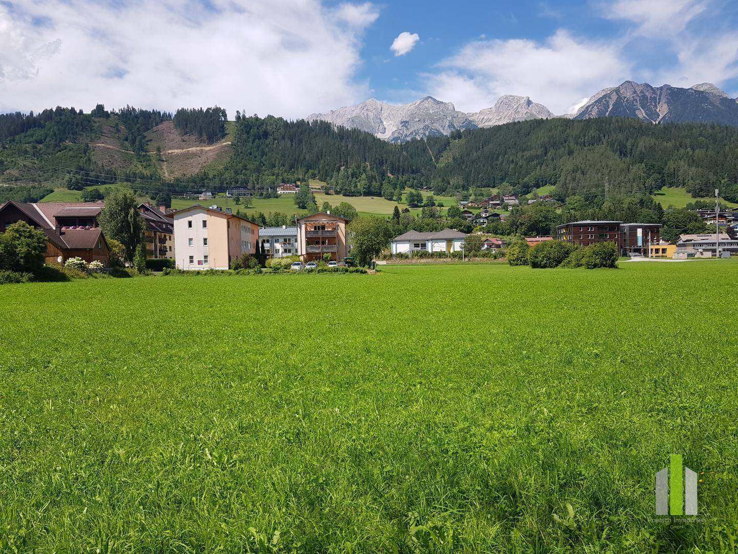 Weitläufiges grünes Feld mit Panoramablick auf die majestätischen Berge und die umliegende Bebauung.