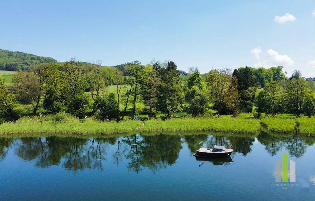 Luftaufnahme eines ruhigen Sees mit einem kleinen Boot, umgeben von bewaldeten Hügeln unter blauem Himmel.