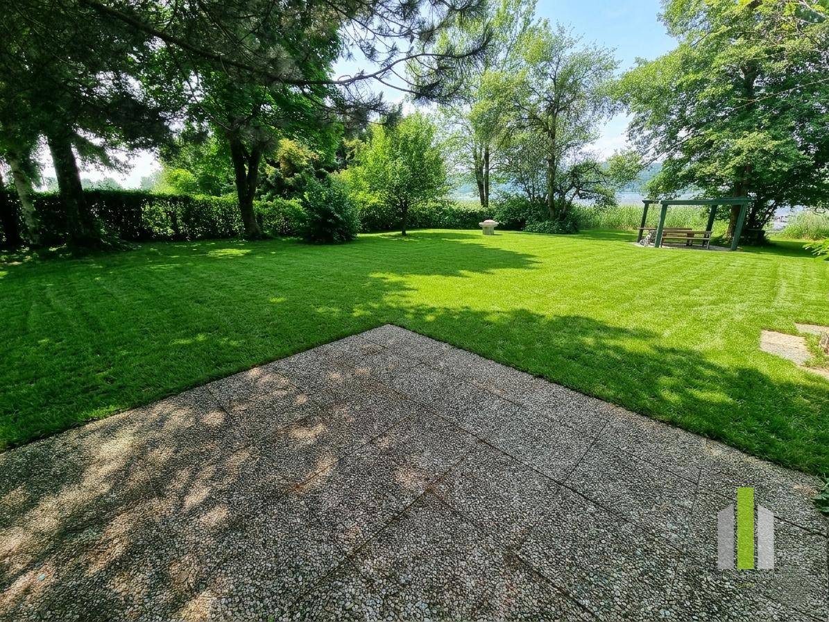 Großer, gepflegter Rasen mit einer gepflasterten Terrasse und weitem Blick auf den See, umgeben von Bäumen.