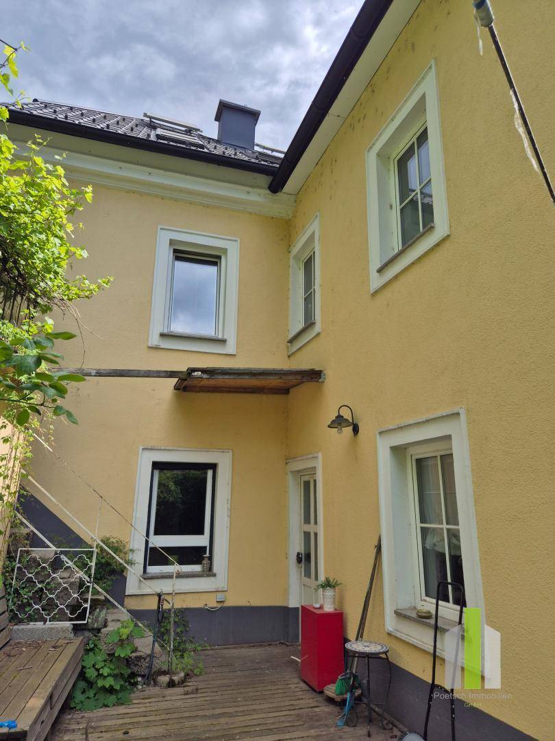 Gelbe Hausfassade mit Holzterrasse und mehreren Fenstern, umgeben von grüner Bepflanzung.