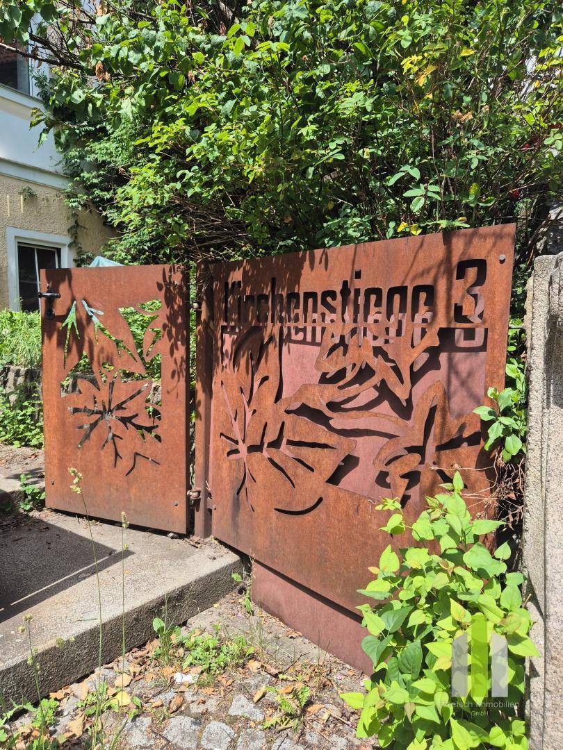 Rostfarbenes Metalltor mit ausgeschnittenen Mustern und Hausnummer, umgeben von grüner Vegetation.