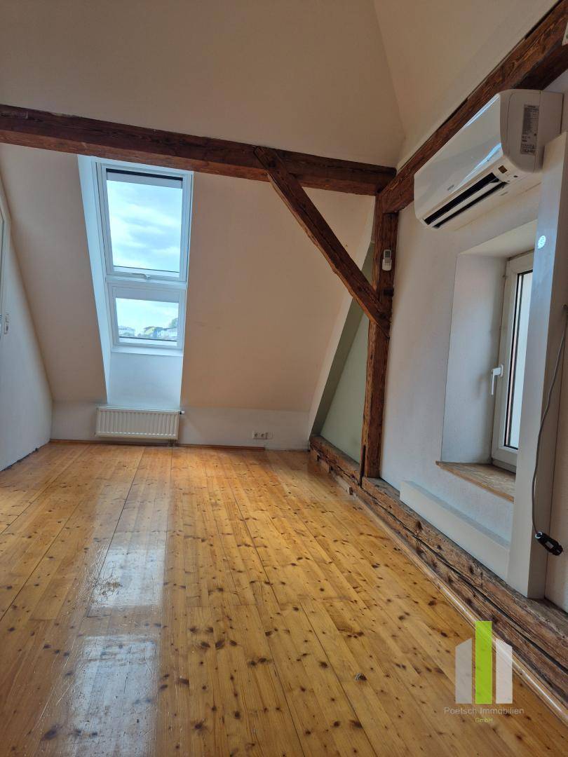 Helles Dachzimmer mit Holzboden, sichtbaren Holzbalken und zwei Dachfenstern für viel Tageslicht.