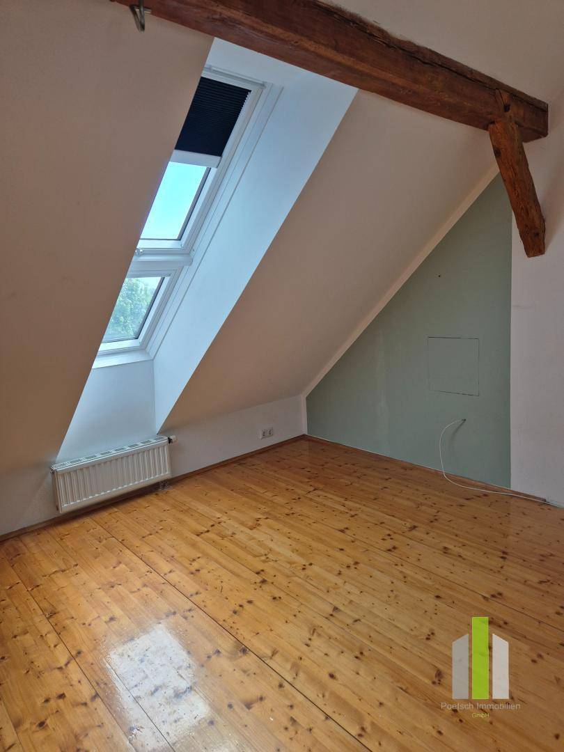 Lichtdurchflutetes Dachzimmer mit Holzboden, Dachschrägen und Heizkörper unter den Dachfenstern.