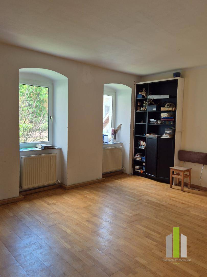 Helles Wohnzimmer mit Holzboden, zwei Fenstern und einem Bücherregal an der Wand.