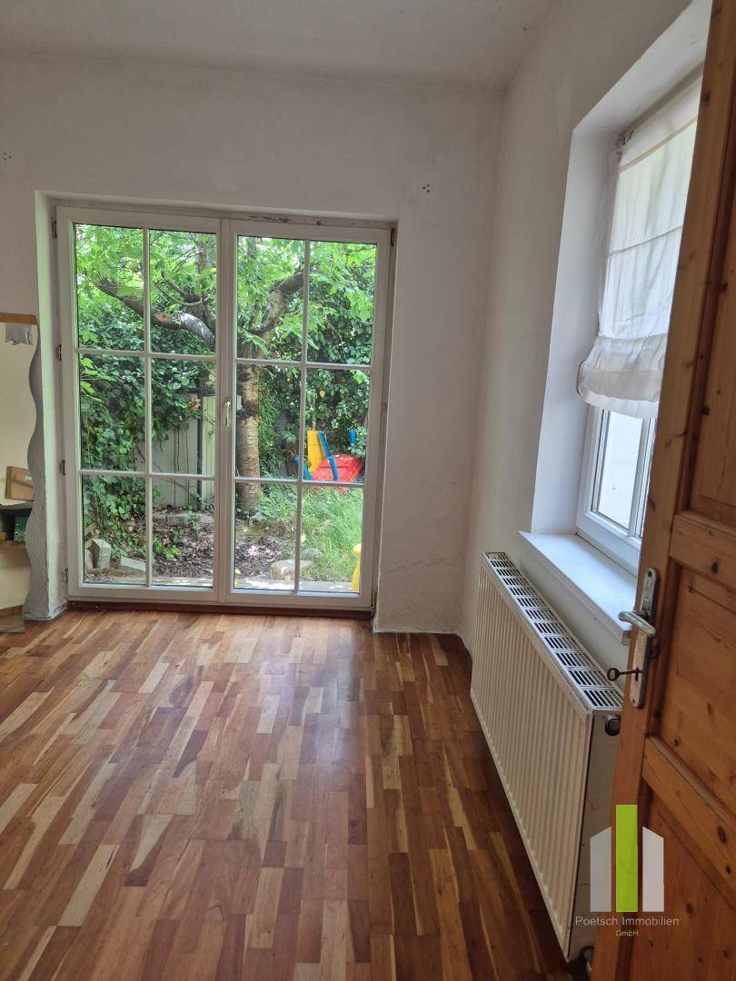 Zimmer mit Holzboden, großer Fenstertür zum Garten und einem Heizkörper unter dem Fenster.