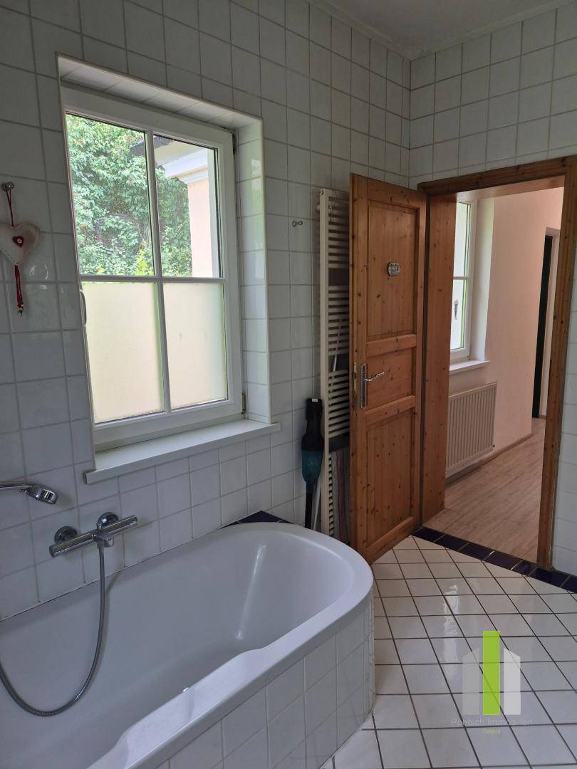 Badezimmer mit Badewanne, weißen Fliesen und einem Fenster, das Tageslicht hereinlässt.
