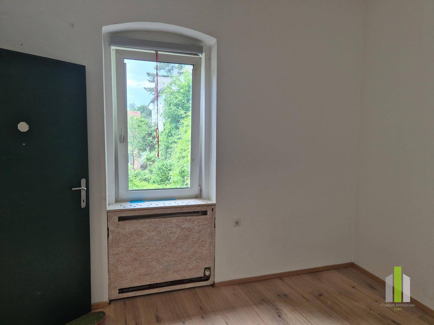 Zimmer mit Holzboden, einem Fenster mit Blick ins Grüne und einer dunklen Tür.