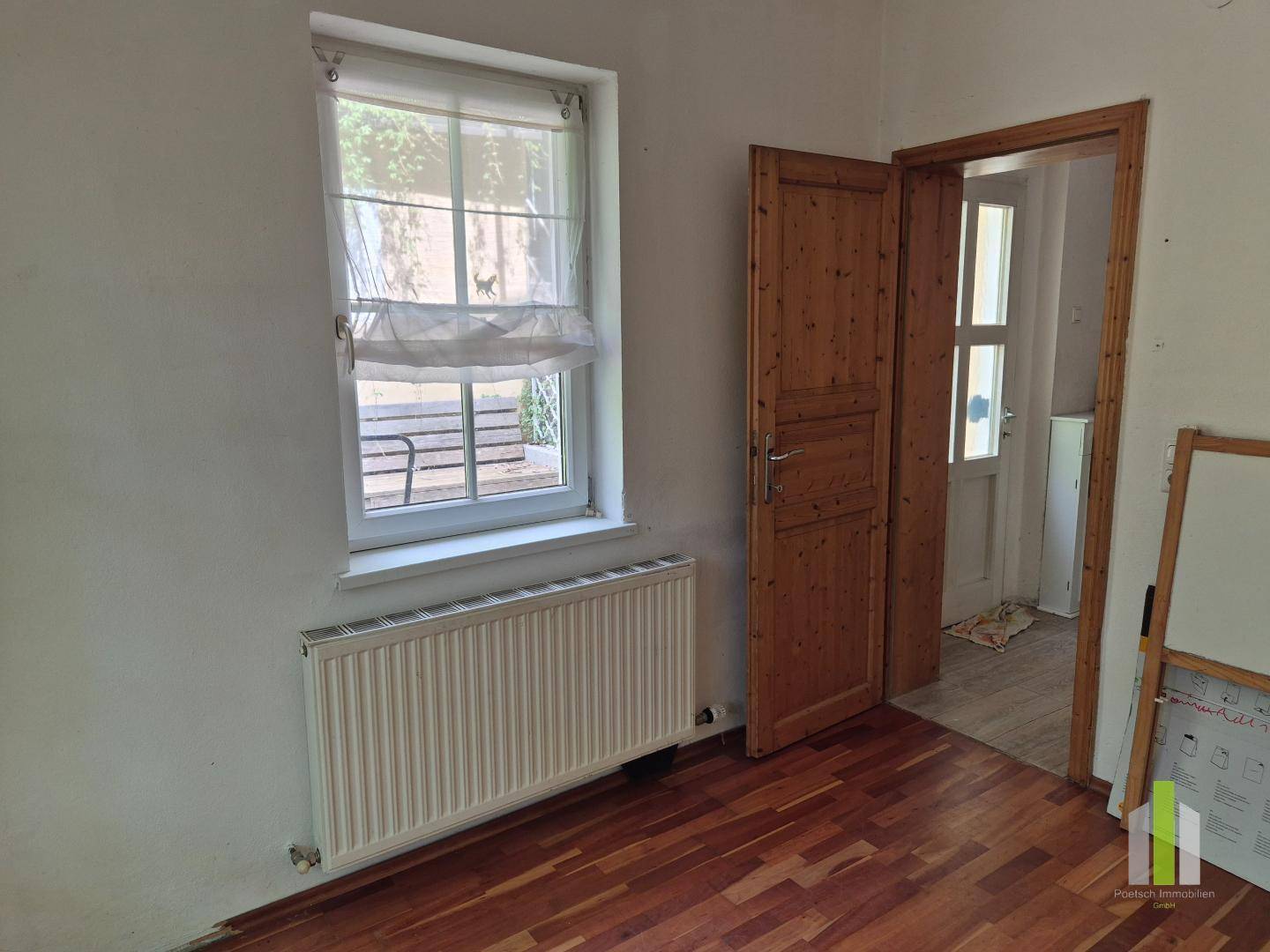 Helles Zimmer mit Holzboden, Fenster und einer Holztür, die in einen anderen Raum führt.