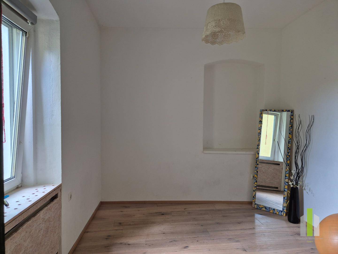 Helles Zimmer mit Holzboden, einem Fenster und einem großen Standspiegel.