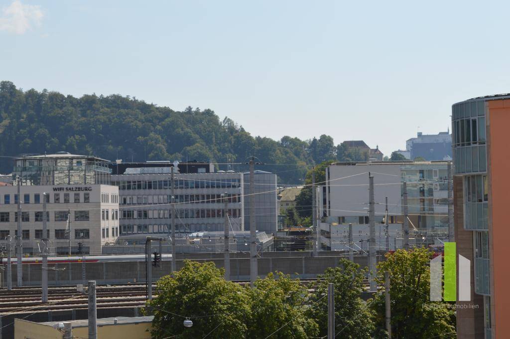 Weitläufiger Blick auf die Stadtlandschaft mit Gebäuden, Bahngleisen und bewaldeten Hügeln im Hintergrund.