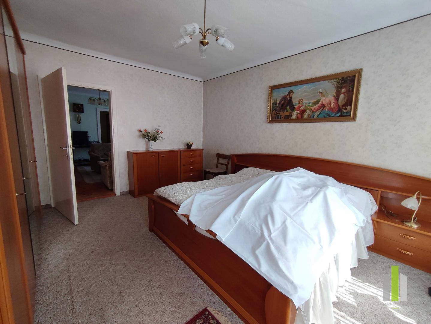 Schlafzimmer mit Doppelbett, Holzmöbeln und Teppichboden, durch eine offene Tür sichtbar.