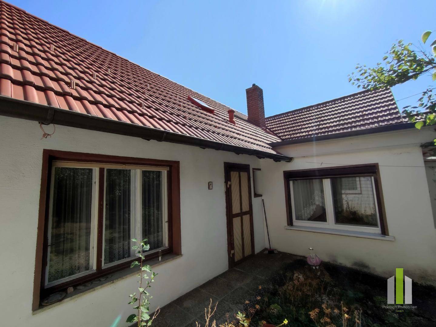 Seitenansicht des Hauses mit rotem Ziegeldach, Fenstern und einem Schornstein unter blauem Himmel.