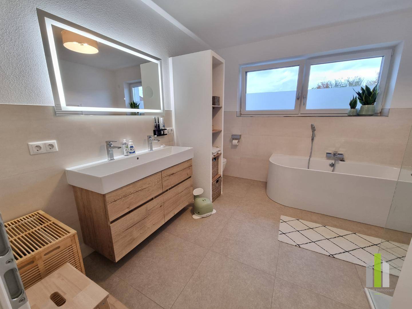 Modernes Badezimmer mit Doppelwaschbecken, beleuchtetem Spiegel und Badewanne am Fenster.