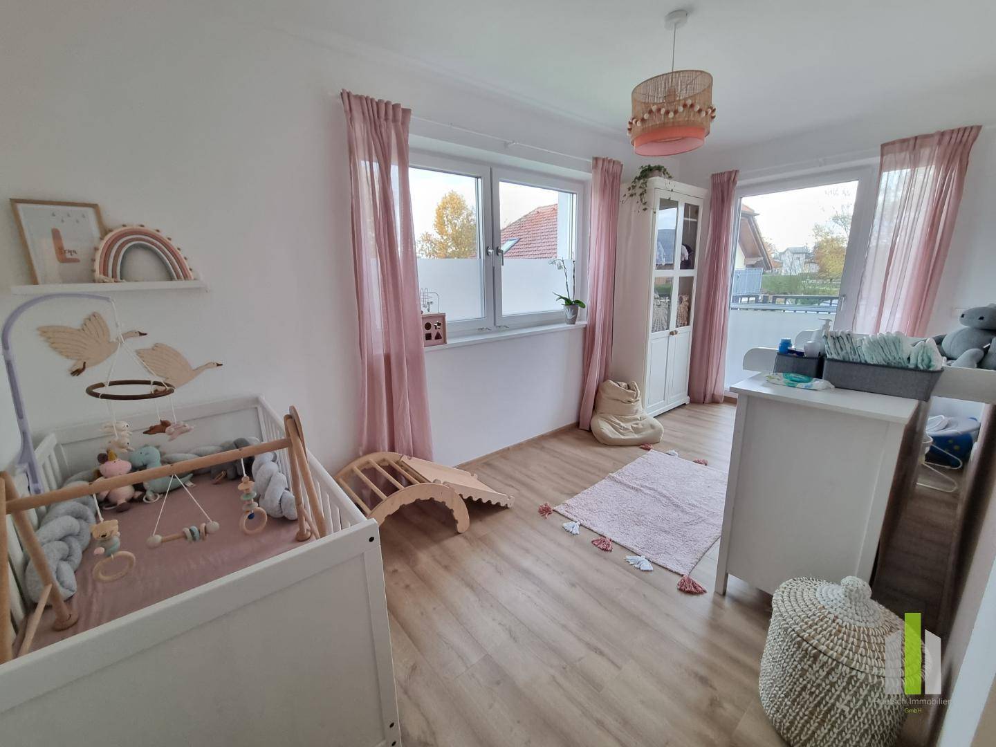 Helles Kinderzimmer mit Babybett, Wickeltisch und rosa Vorhängen, ausgestattet mit Holzboden.