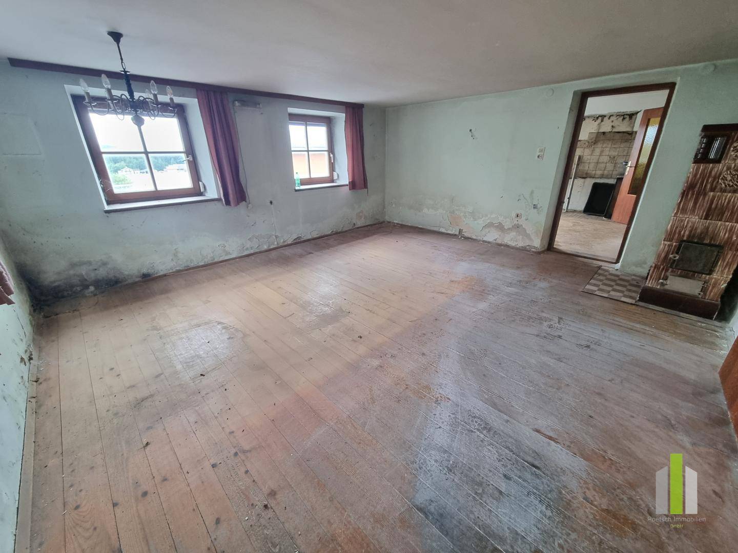Geräumiges Wohnzimmer mit Holzdielenboden, zwei Fenstern und einem traditionellen Kachelofen.