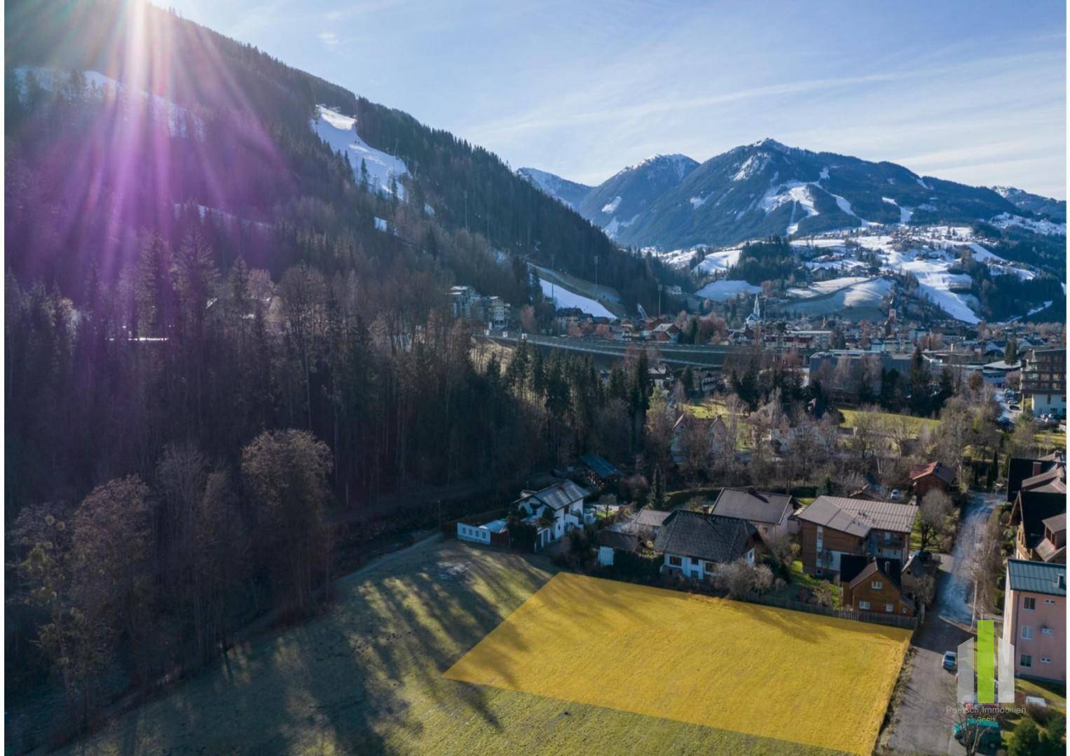 Sonnenbeschienene Luftaufnahme eines Baugrundstücks mit gelb markierter Fläche, umgeben von Wald und Bergen.