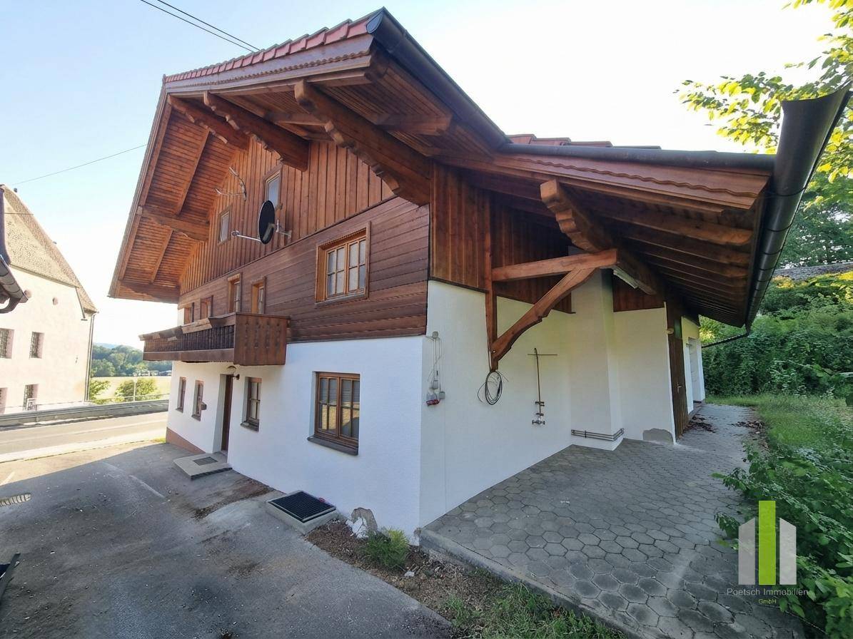 Traditionelles Holzhaus mit weißer Fassade, Holzbalkon und gepflasterter Zufahrt unter blauem Himmel.