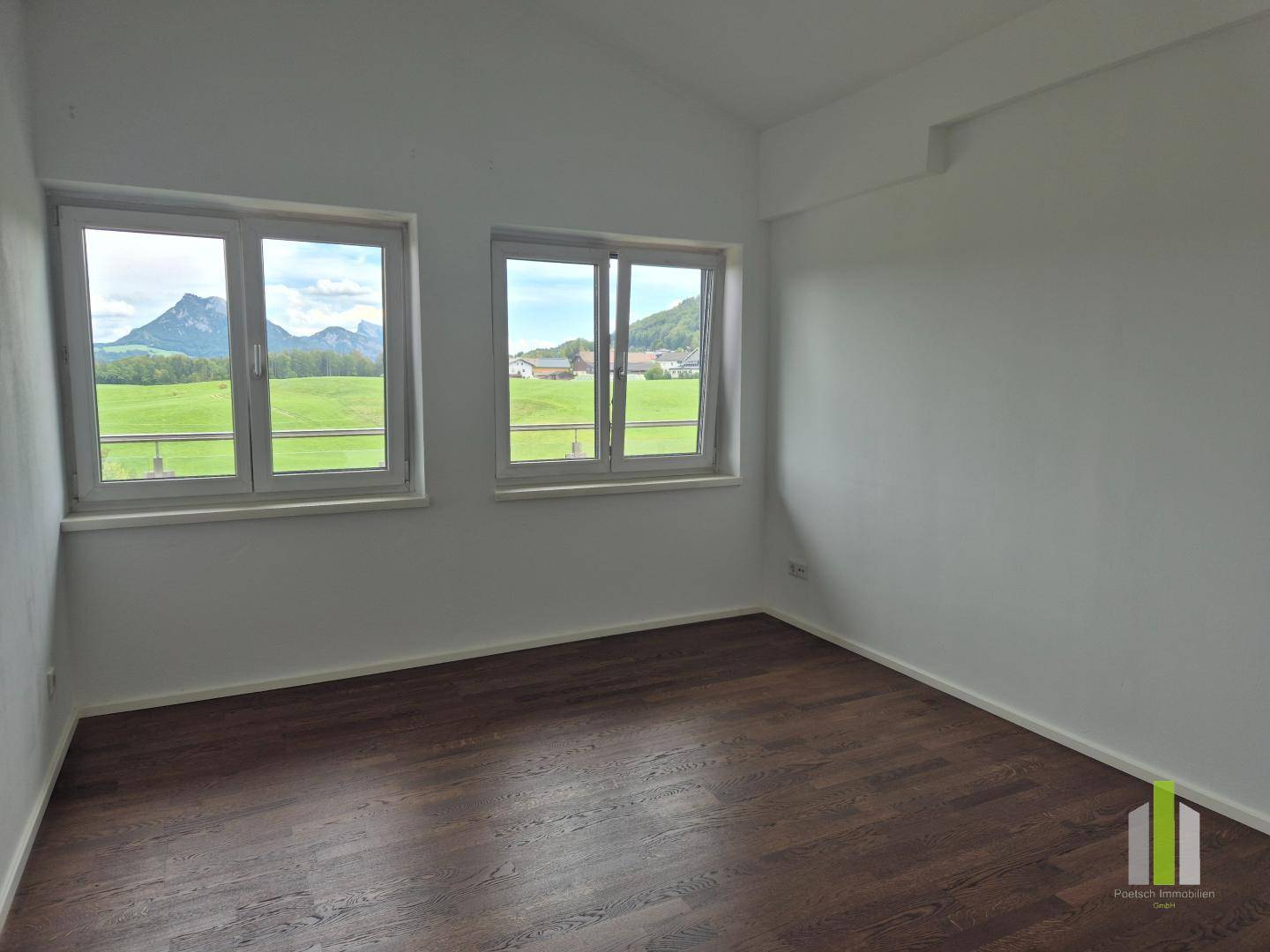 Helles Zimmer mit zwei Fenstern, dunklem Holzboden und weitem Blick auf grüne Felder und Berge.