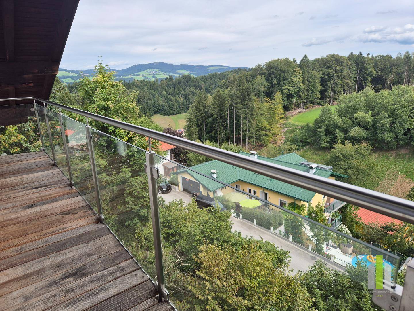 Geräumiger Balkon mit Holzboden und Glasgeländer, bietet einen Panoramablick auf bewaldete Hügel und Häuser.