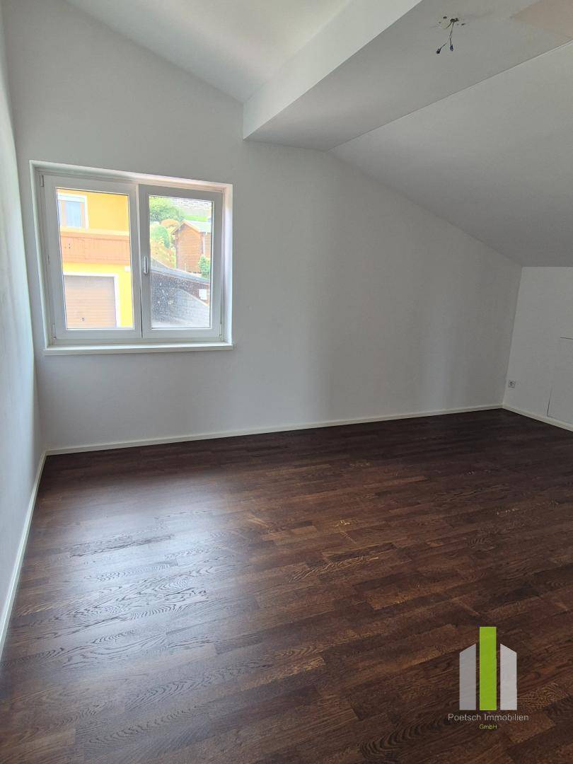 Geräumiges Zimmer mit Dachschräge, dunklem Holzboden und einem Fenster mit Blick auf die Nachbarschaft.