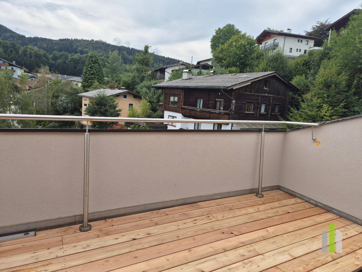 Kleiner Balkon mit Holzboden und Edelstahlgeländer, umgeben von grünen Hügeln und Häusern.