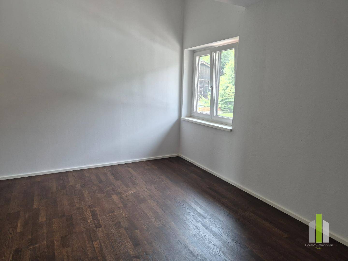 Schlichtes Zimmer mit dunklem Holzboden und einem Fenster, das viel natürliches Licht hereinlässt.