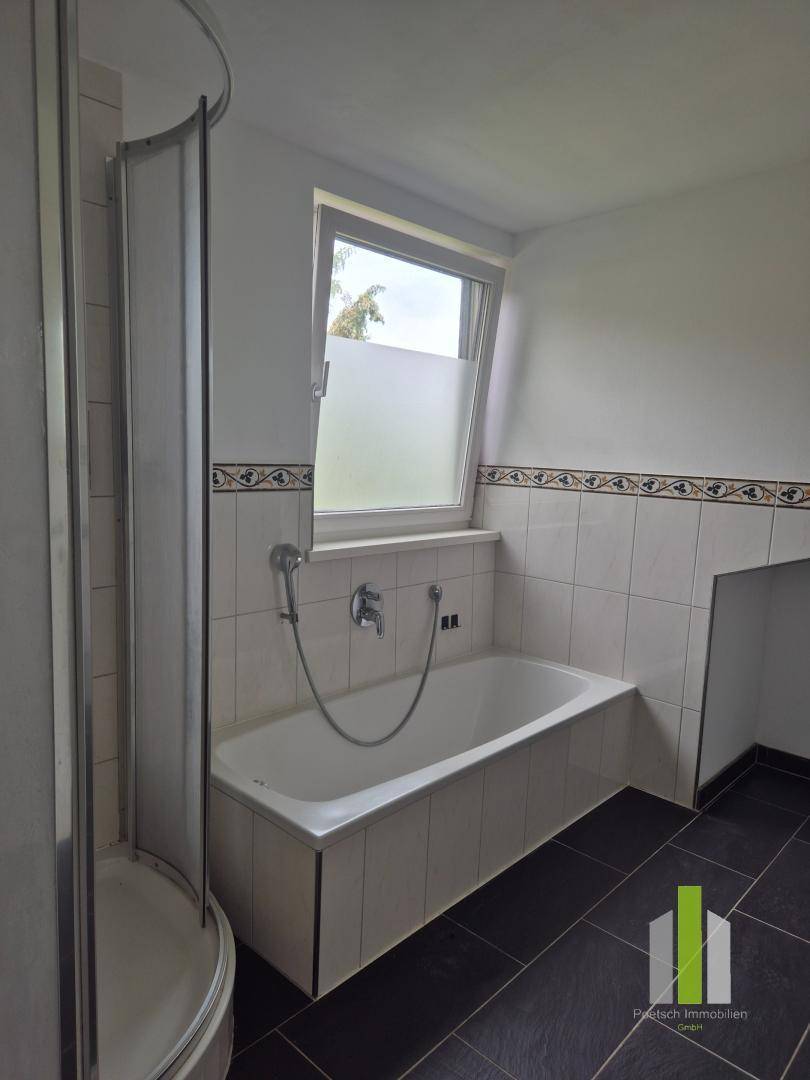Badezimmer mit Badewanne, Duschkabine und Fenster, ausgestattet mit dunklen Bodenfliesen.