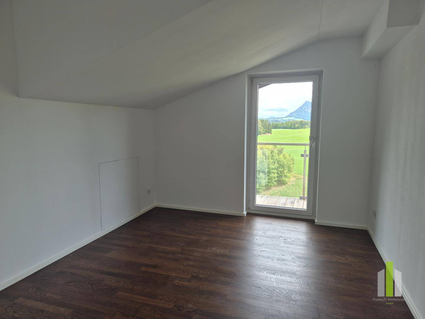 Helles Zimmer mit Dachschräge, dunklem Holzboden und Balkonzugang mit Bergblick.
