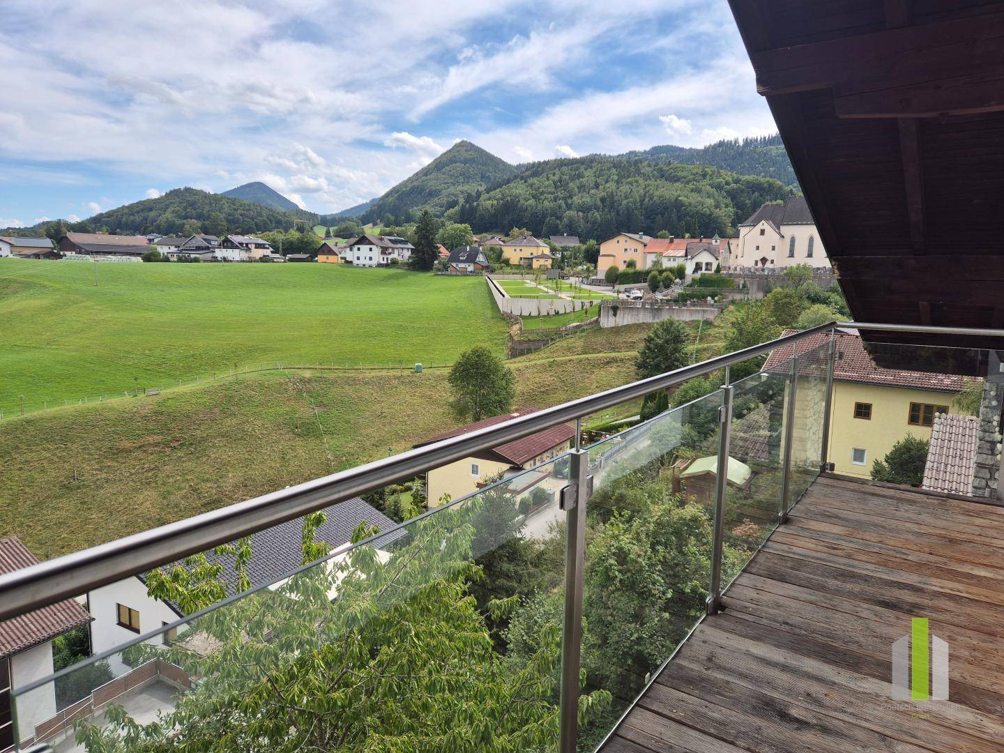 Großer Balkon mit Holzboden und Glasgeländer, bietet einen weiten Blick auf die Landschaft und ein Dorf.