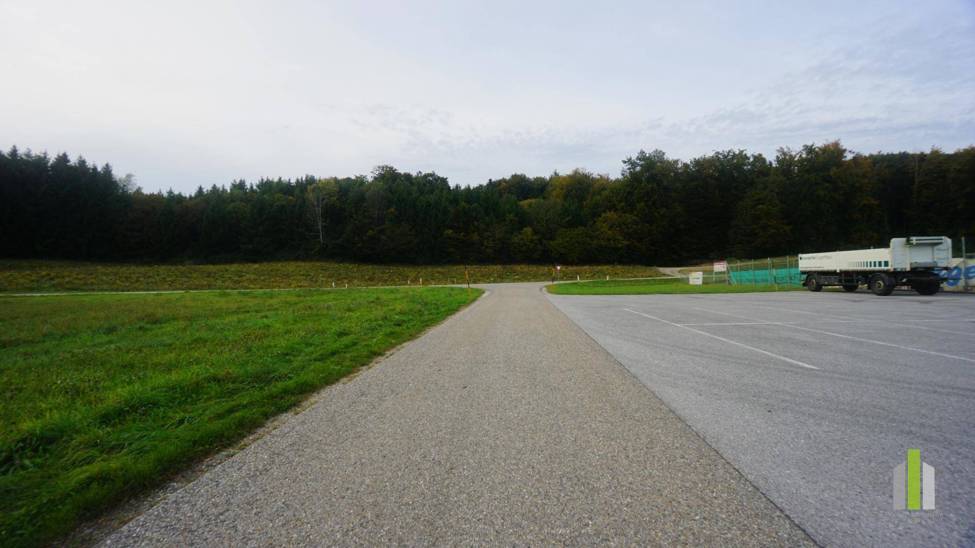 Geschotterter Weg führt durch grüne Wiesen zu einem Waldrand, mit einem großen Parkplatz rechts.