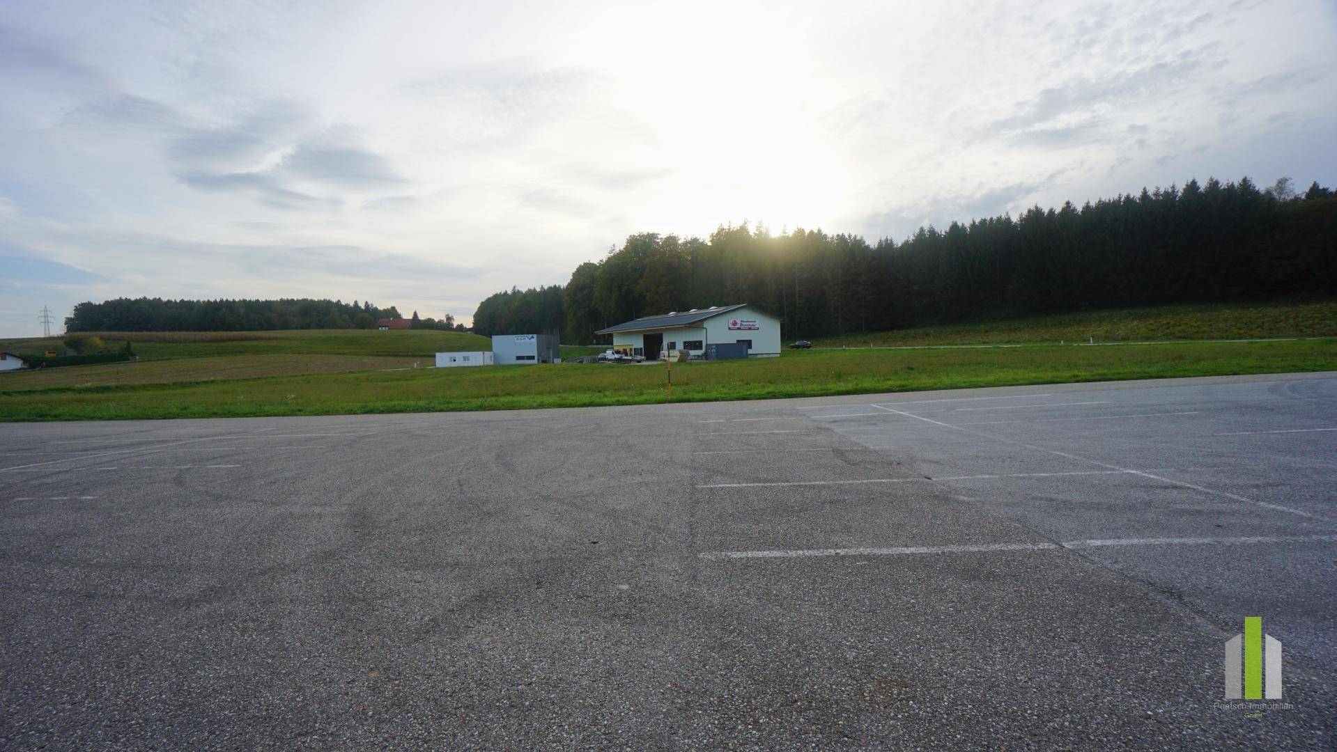 Großer asphaltierter Parkplatz im Vordergrund, dahinter grüne Felder, Gebäude und Wald bei Sonnenschein.