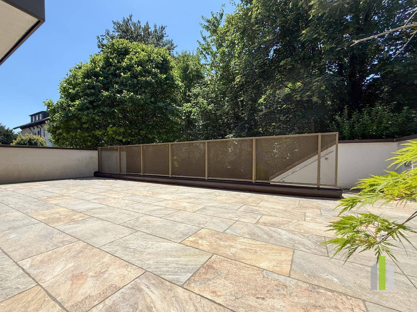 Weitläufige, geflieste Terrasse umgeben von üppigem Grün und hohen Bäumen im Hintergrund.