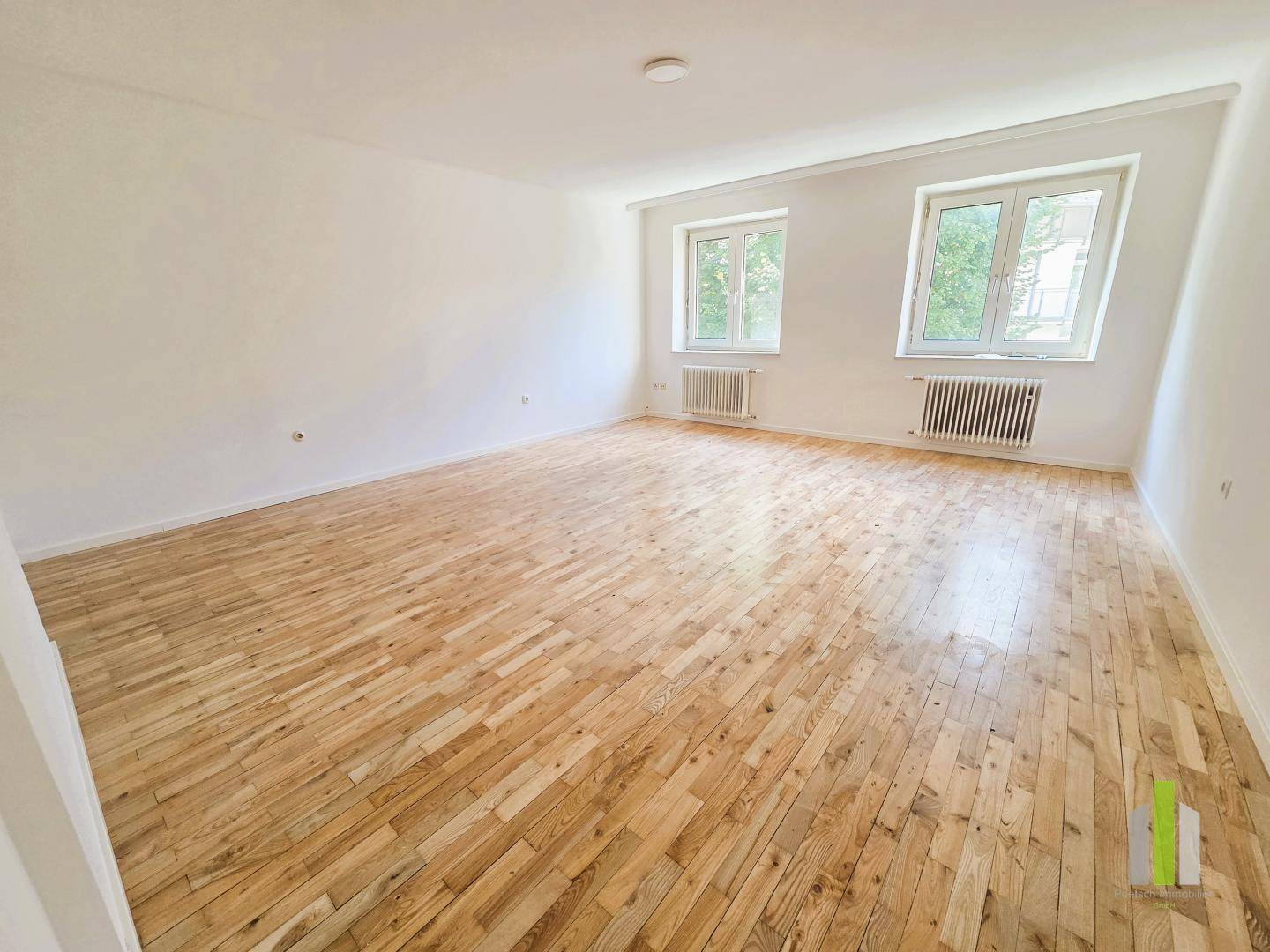 Helles, geräumiges Zimmer mit Holzdielenboden und zwei großen Fenstern, ideal für ein Wohnzimmer.
