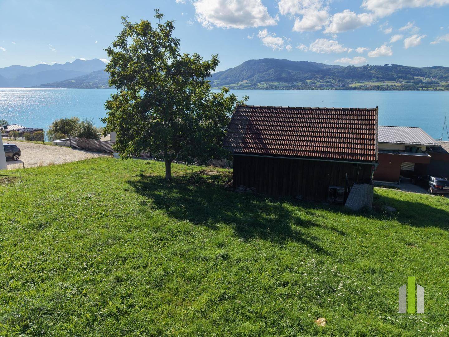 Grüne Wiese mit einem kleinen Holzschuppen und Baum, im Hintergrund der See.