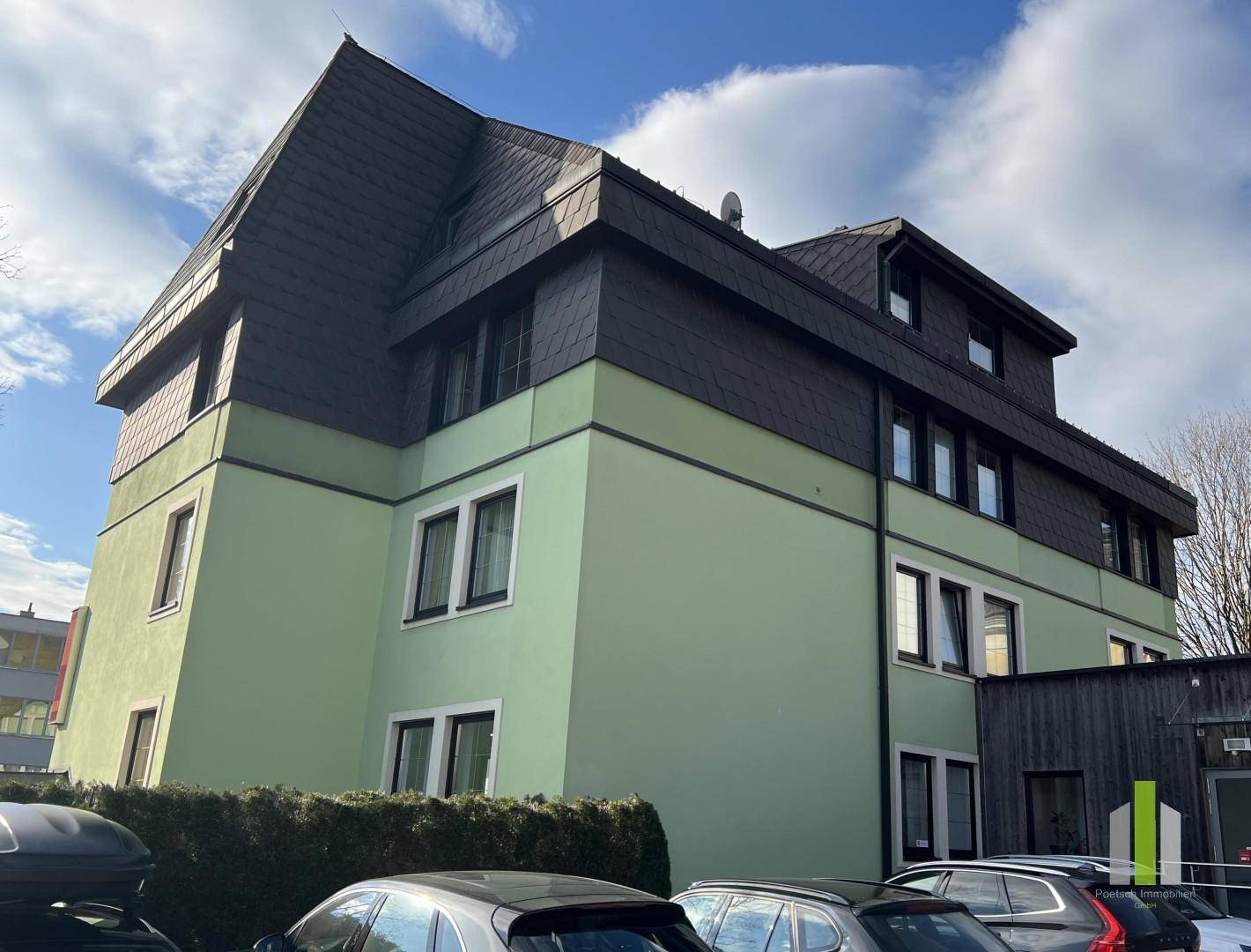 Seitenansicht eines grünen Gebäudes mit dunklem Dach und geparkten Autos davor.