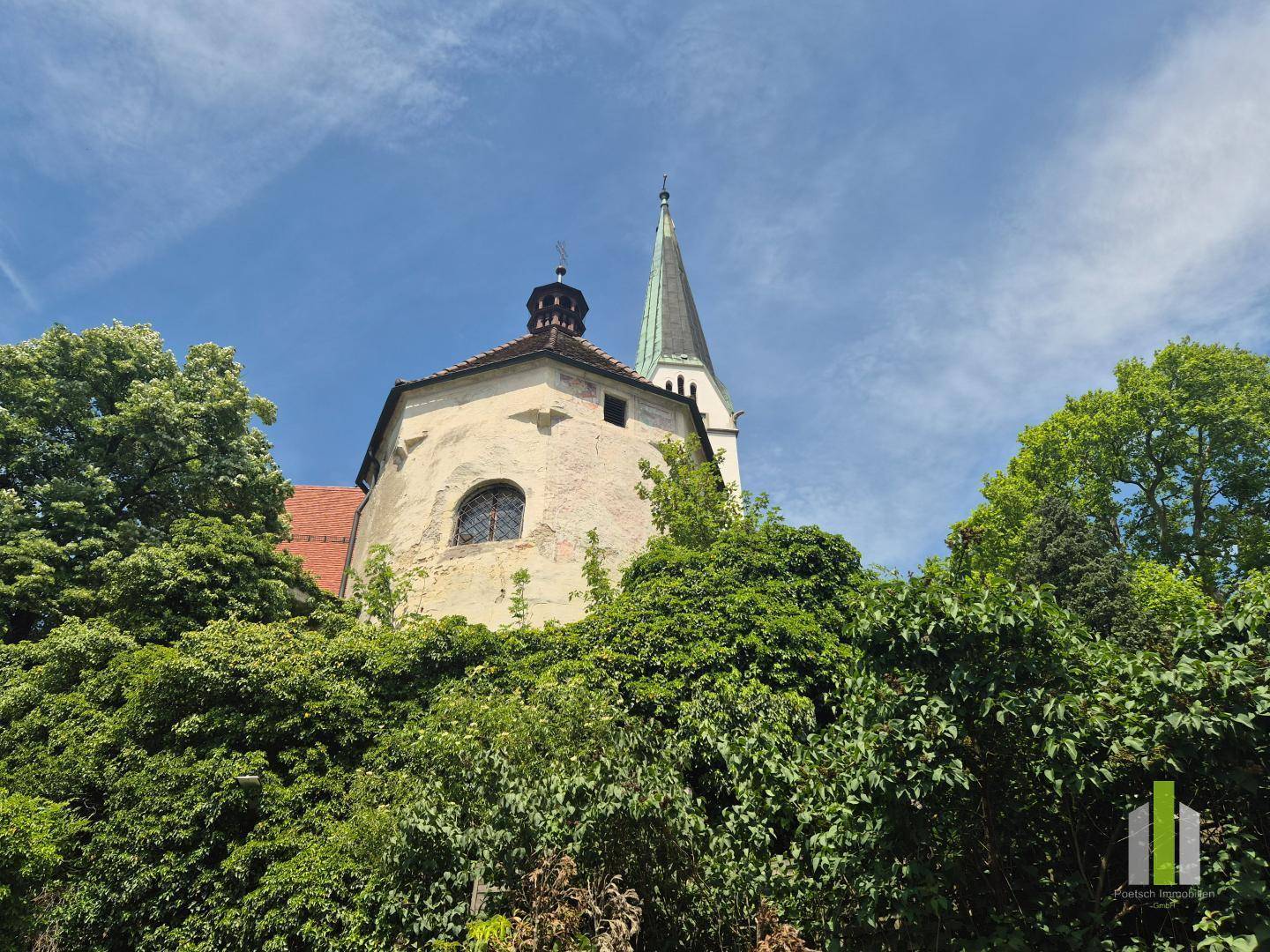 Historischer Kirchturm und Gebäude, umgeben von üppigem Grün unter blauem Himmel.