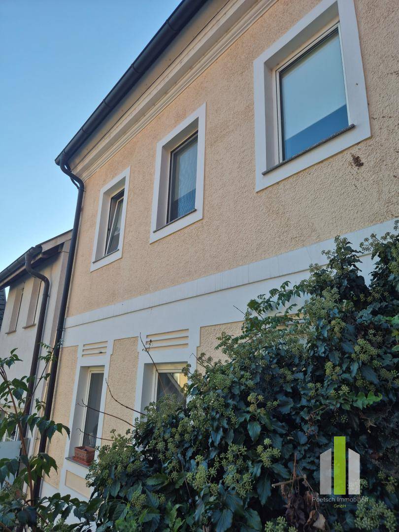 Seitenansicht eines Gebäudes mit beiger Fassade und grüner Bepflanzung im Vordergrund.