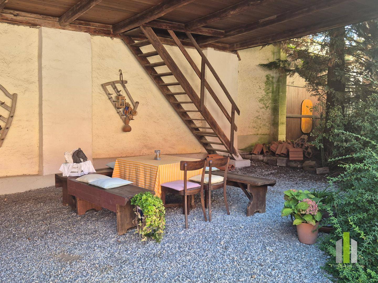 Rustikaler Essbereich im überdachten Innenhof mit Holztisch, Bänken und einer Holztreppe. 