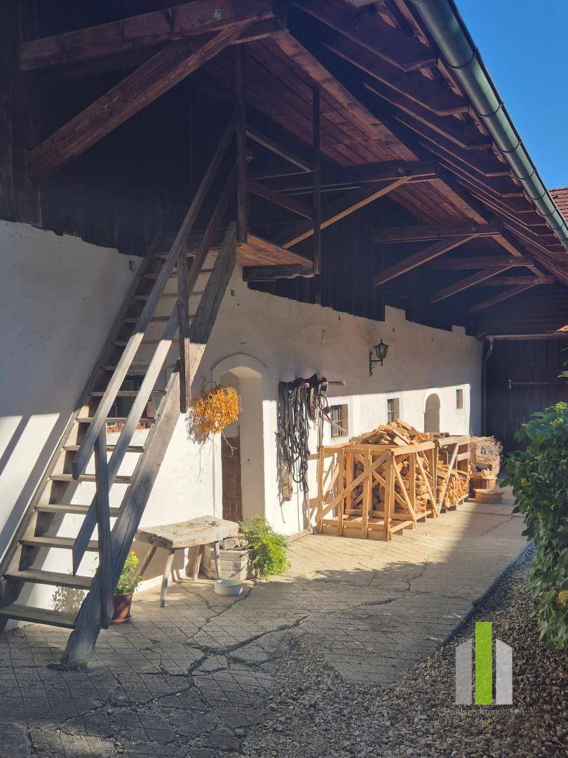 Überdachter Außenbereich mit Holztreppe, Brennholzstapeln und einem gepflasterten Weg. 