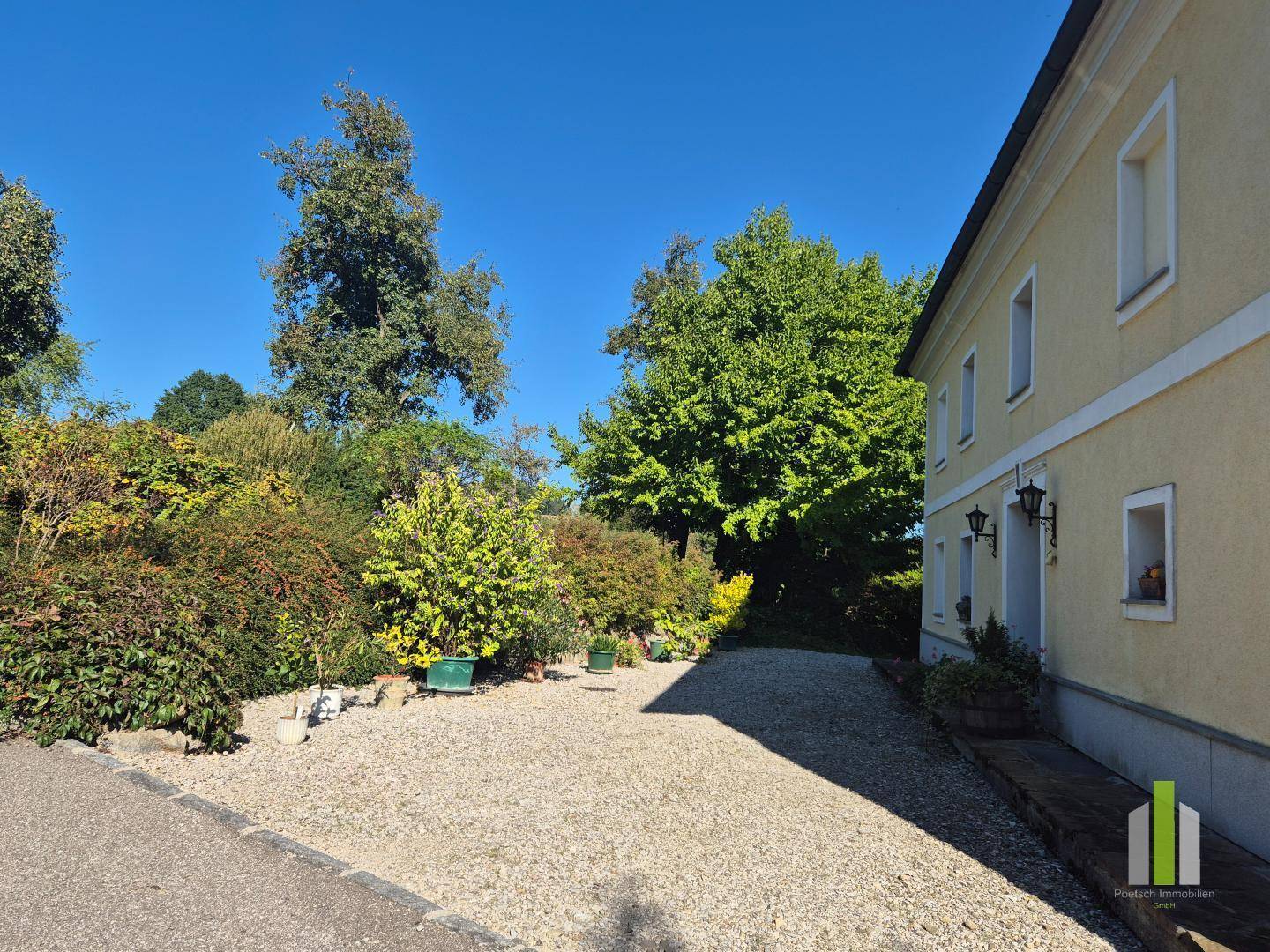 Seitenansicht des Hauses mit Kiesauffahrt und üppiger grüner Bepflanzung unter sonnigem Himmel. 