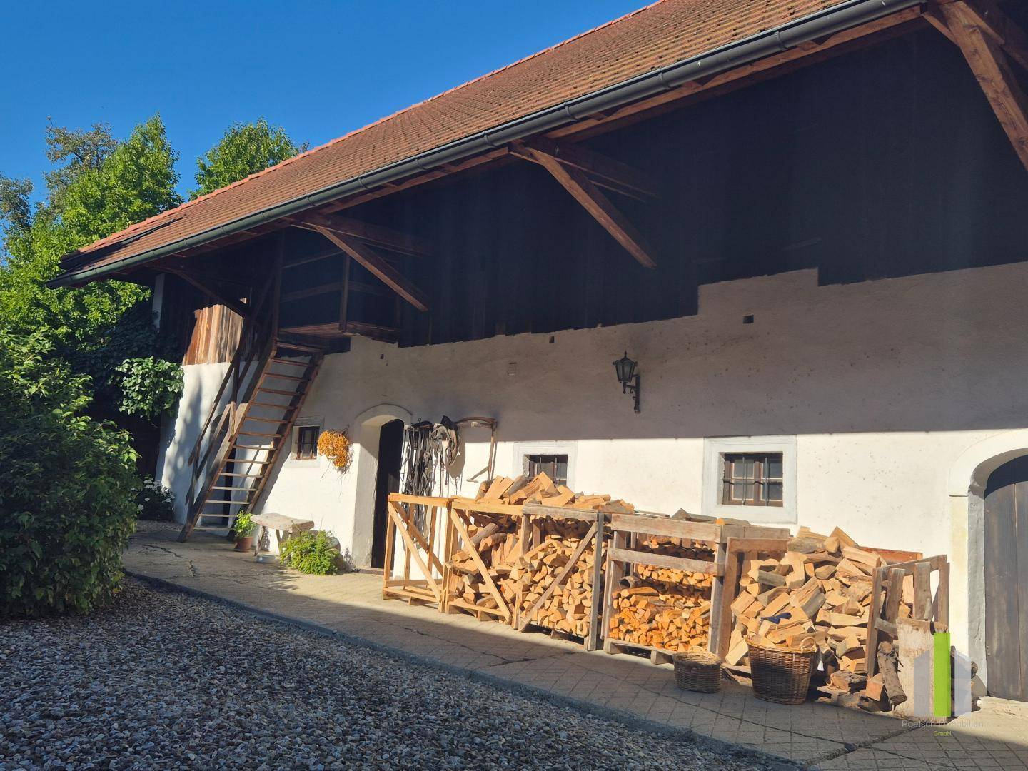 Rustikales Bauernhaus mit Holzlager und Außentreppe unter blauem Himmel, traditioneller Stil.