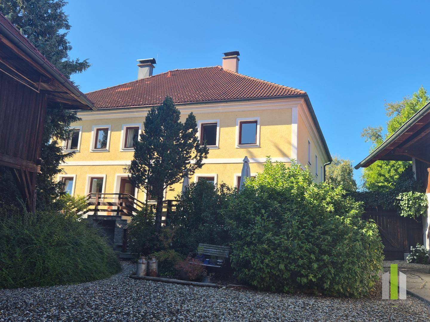 Gesamtansicht des gelben Hauses umgeben von grüner Bepflanzung und einem Kiesbereich. 
