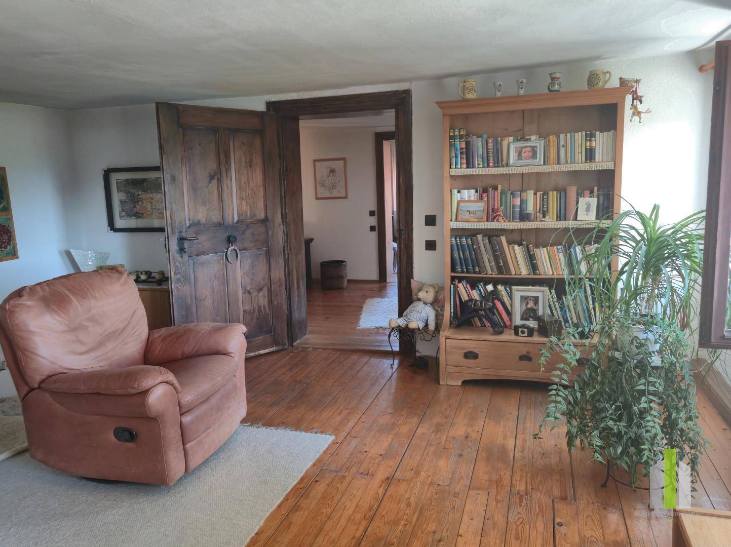 Wohnzimmer mit Ledersessel, Holzboden und Bücherregal, das zu einem weiteren Raum führt.