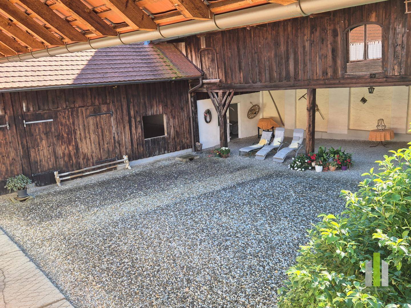 Kiesiger Innenhof mit Liegestühlen unter einem überdachten Bereich und angrenzender Scheune. 