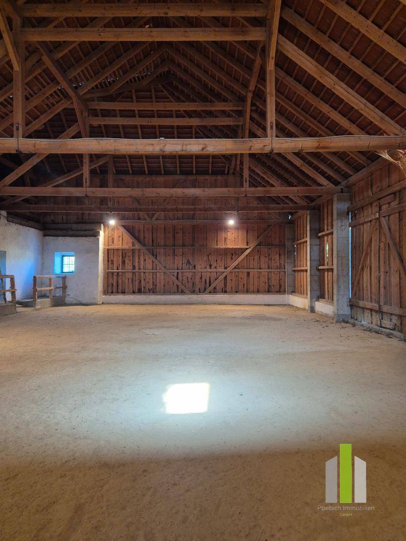 Weitläufiger Innenraum einer Scheune mit sichtbaren Holzbalken und natürlichem Lichteinfall durch ein kleines Fenster. 